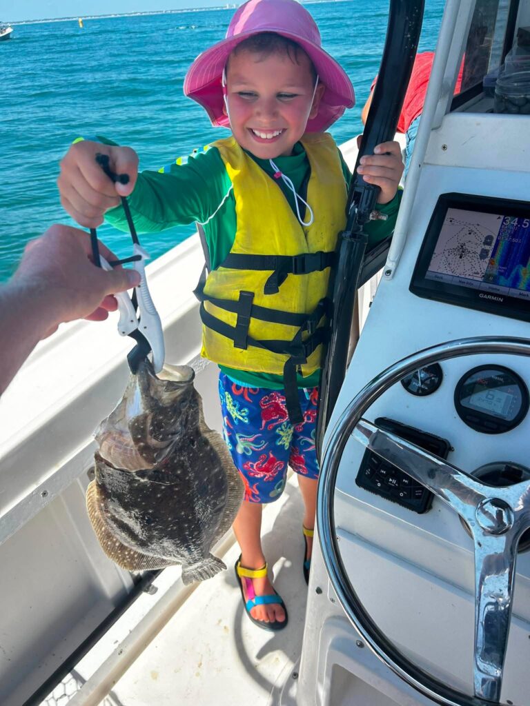 Child in a life jacket and pink sun hat holding a flounder on a fishing boat