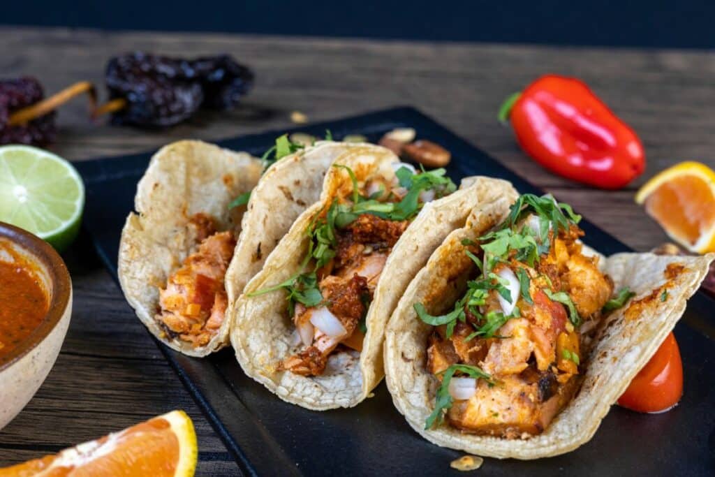 Three fish tacos topped with cilantro and onion beside lime and orange wedges